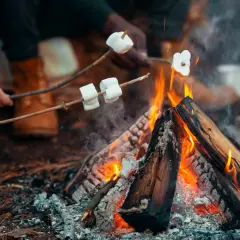 Ręce trzymają patyki z piankami na ognisko. Dym unosi się nad płomieniami, a w tle widać buty.