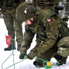 Saper w wojsku przygotowuje teren w zimowych warunkach. Żołnierz w kamuflażu pochyla się nad zielonymi przewodami, pracując ostrożnie.