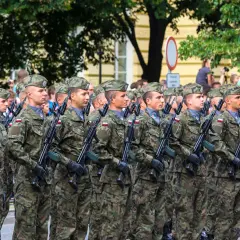 Żołnierze w mundurach i beretach stoją w zwartym szyku, trzymając broń. W tle widać drzewa i budynki, a także flagę. To widok stanowisk wojskowych podczas parady.
