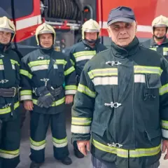 Starszy strażak OSP, w towarzystwie kolegów, gotowy do akcji. W tle wóz strażacki.