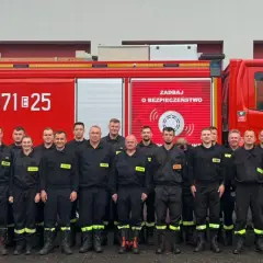 Grupa strażaków w mundurach, obok czerwonego wozu strażackiego. Dowódca sekcji OSP stoi w pierwszym rzędzie, w jasnym mundurze.