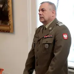 Generał w mundurze, z orłem na piersi, stoi w sali, gdzie odbywa się spotkanie korpusu generałów.