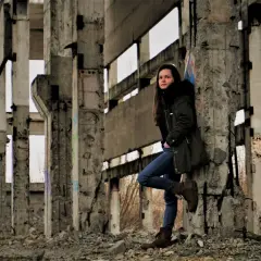 Młoda kobieta w ciemnej kurtce i jeansach opiera się o betonowy filar w zrujnowanym budynku. To miejsce idealne do chodzenia po opuszczonych budynkach, pełne surowego piękna i historii.