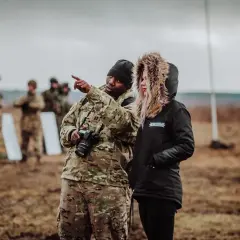 Żołnierz w mundurze pokazuje coś kobiecie w kurtce z futrzanym kapturem. W tle inni żołnierze. Pytanie "w jakim wieku do wojska" może być związane z tym szkoleniem.