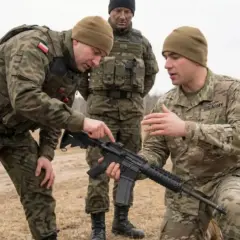 Żołnierze, w tym polscy i amerykańscy, ćwiczą obsługę broni. Dwóch żołnierzy w wojsku omawia szczegóły karabinu.