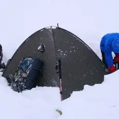 Osoba w niebieskiej kurtce i czerwonych rękawiczkach przygotowuje namiot w śniegu. Obok leżą plecaki i kijki trekkingowe, które pomagają dobrać kijki trekkingowe do wzrostu.