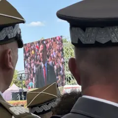 Dwóch mężczyzn w mundurach, jeden z nich ma jedną gwiazdkę na pagonie, ogląda przemówienie na telebimie.