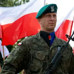 Żołnierz w zielonym berku i mundurze, z karabinem na ramieniu, na tle polskich flag. Widać jego stopnie wojskowe i nazwisko.