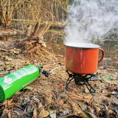 Kuchenka turystyczna z gazem i parującym kubkiem nad wodą. Różne rodzaje kuchenek pozwalają na gotowanie w plenerze.