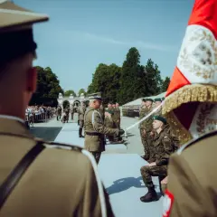 Podczas uroczystości wojskowej, oficer wręcza szablę klęczącemu podporucznikowi. W tle stoją żołnierze i widzowie.