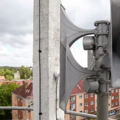 Syrena alarmowa na budynku, gotowa do ogłoszenia, jak brzmi alarm bombowy w Polsce. W tle zabudowania miejskie.