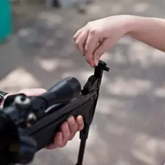 Dziecko uczy się obsługi **broni pneumatycznej** pod okiem dorosłego. Widać lunetę i mechanizm ładowania.
