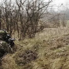 Gracz w kamuflażu leży w trawie, celując z repliki ASG. Czy można strzelać ASG w lesie? To zależy od przepisów.