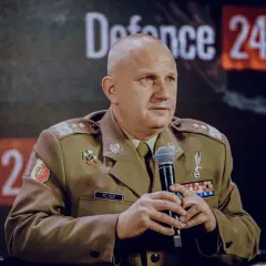 Generał Klisz w mundurze, z mikrofonem w dłoniach, siedzi przed ekranem z napisem "Defence 24 DAYS".