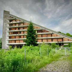 Opuszczony budynek o trójkątnym kształcie, z oknami zabitymi deskami, otoczony bujną zielenią. Idealne urbex miejsca dla poszukiwaczy przygód.