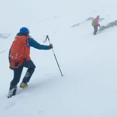 Dwie osoby wspinają się w góry w trudnych warunkach śniegowych. Mają na sobie kaski, plecaki i raki.