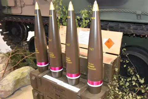 Cztery pociski amunicji artyleryjskiej 155 mm, każdy z oznaczeniem TNT, stoją obok drewnianych skrzyń i gąsienic czołgu.