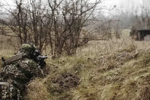 Gracz w kamuflażu leży w trawie, celując z repliki ASG. Czy można strzelać ASG w lesie? To zależy od przepisów.
