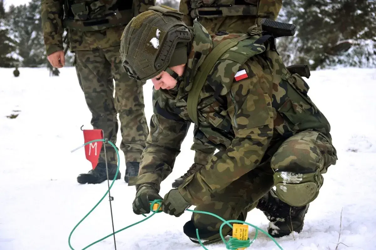 Saper w wojsku przygotowuje teren w zimowych warunkach. Żołnierz w kamuflażu pochyla się nad zielonymi przewodami, pracując ostrożnie.