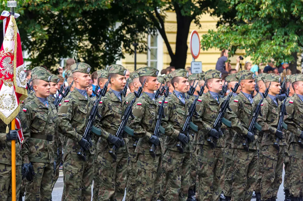 Żołnierze w mundurach i beretach stoją w zwartym szyku, trzymając broń. W tle widać drzewa i budynki, a także flagę. To widok stanowisk wojskowych podczas parady.