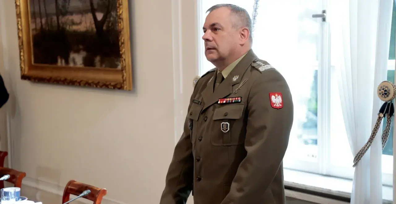 Generał w mundurze, z orłem na piersi, stoi w sali, gdzie odbywa się spotkanie korpusu generałów.