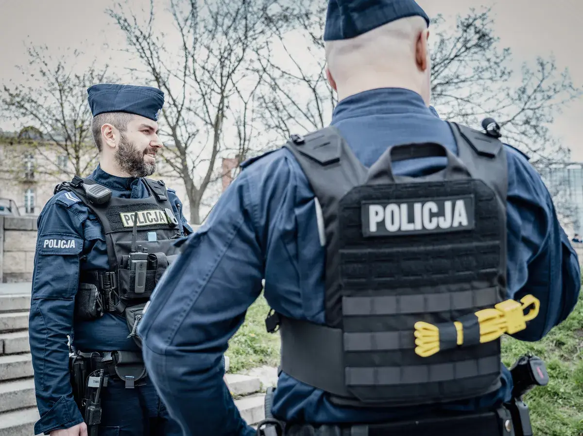 Dwóch policjantów w mundurach i kamizelkach taktycznych. Widoczne wyposażenie policjanta: radiostacja, kabura na broń, pas z ładownicami.