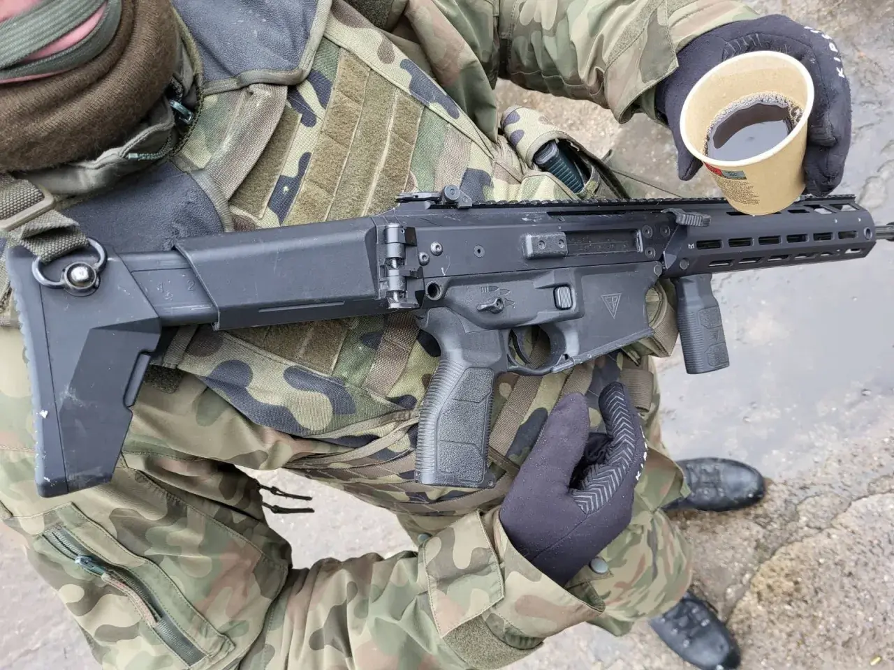 Żołnierz w kamuflażu trzyma karabin i kubek kawy. Czy to broń kolekcjonerska czy sportowa?