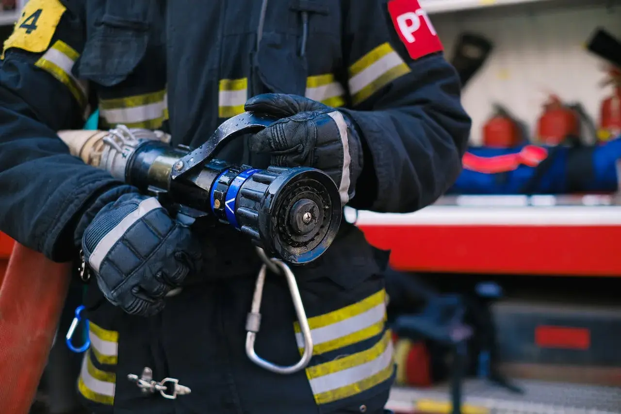 Strażak w rękawicach trzyma prądownicę. Zastanawiasz się, jakie są stopnie w PSP po ilu latach?