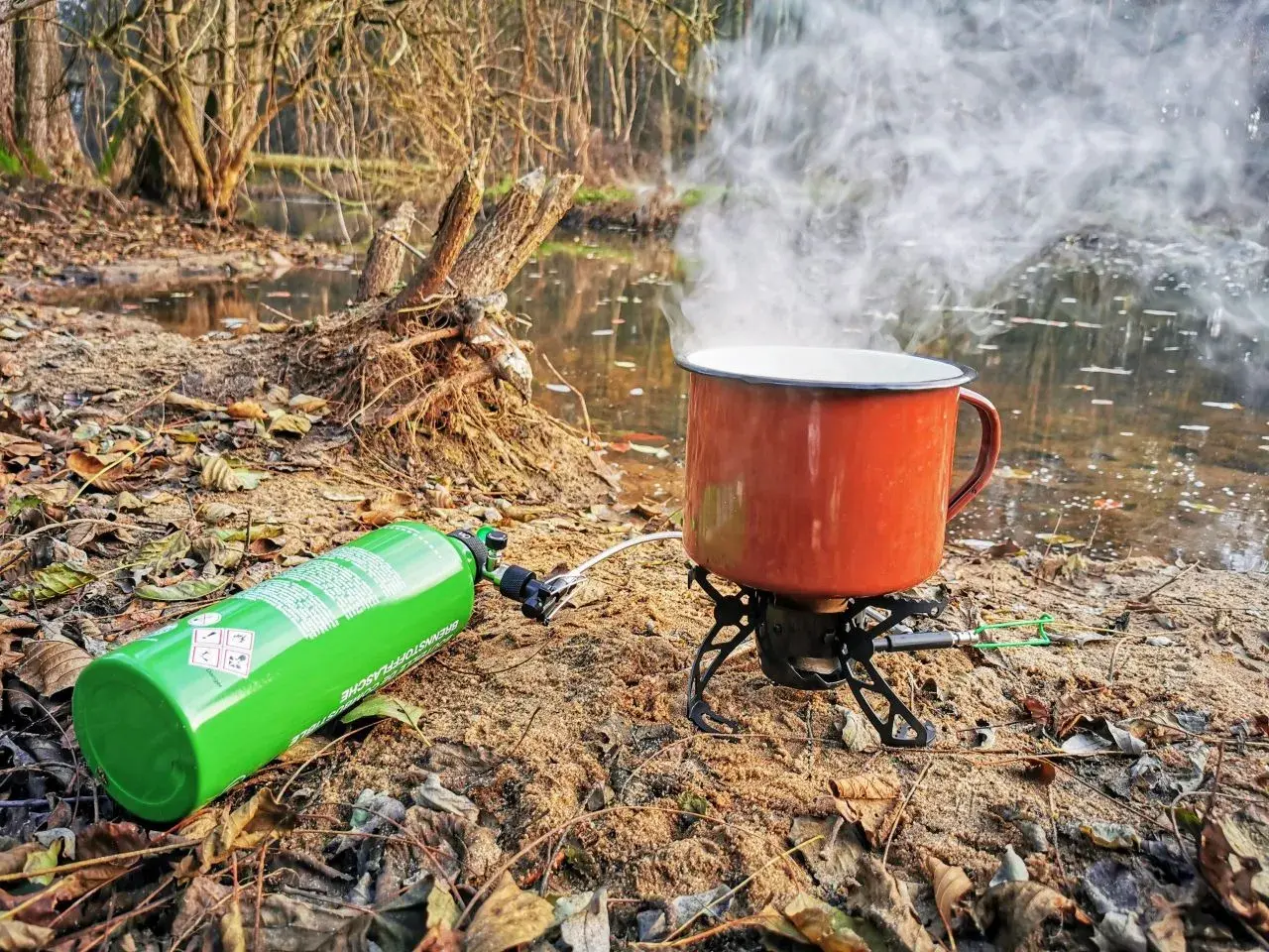Kuchenka turystyczna z gazem i parującym kubkiem nad wodą. Różne rodzaje kuchenek pozwalają na gotowanie w plenerze.