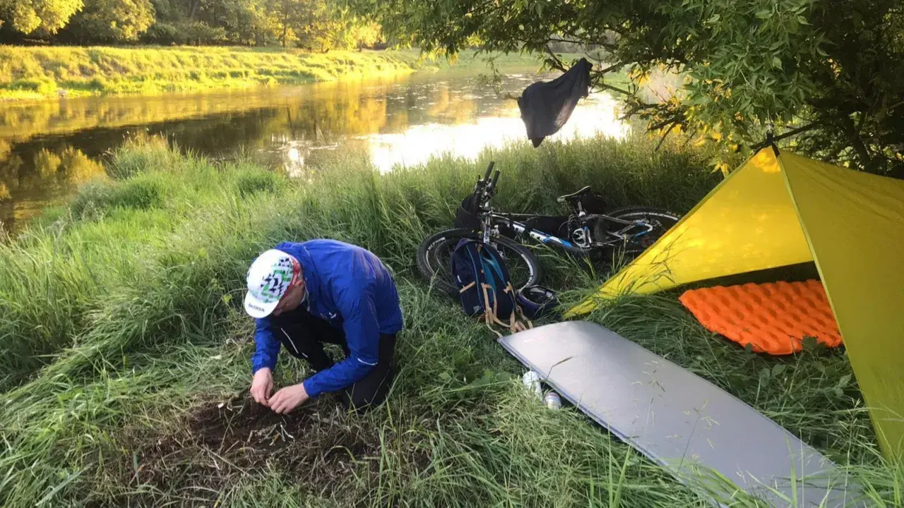 Biwak nad rzeką: rozłożony żółty namiot, materac, rower i mężczyzna przygotowujący ognisko. To idealny materiał na namiot i nocleg w plenerze.