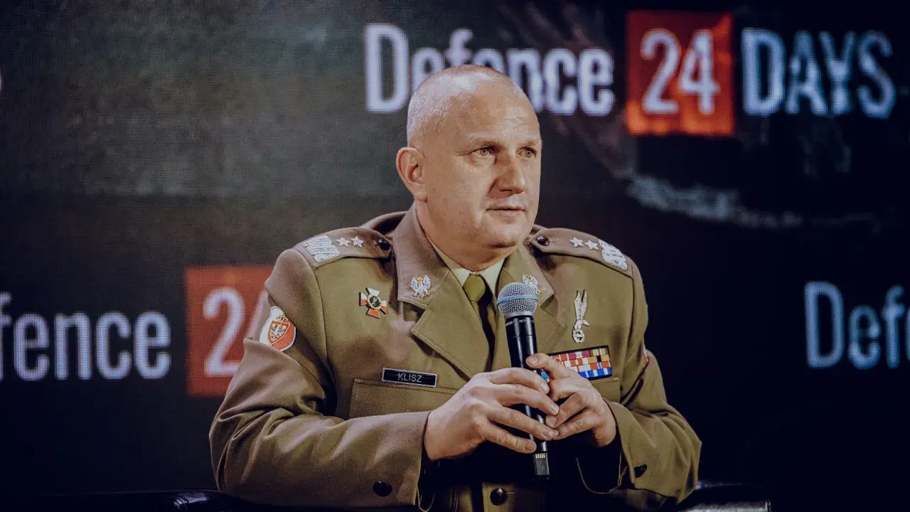 Generał Klisz w mundurze, z mikrofonem w dłoniach, siedzi przed ekranem z napisem "Defence 24 DAYS".