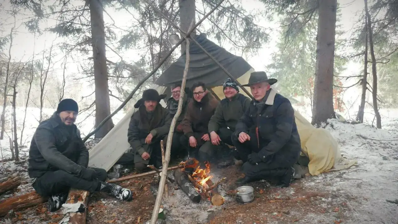 Grupa mężczyzn siedzi przy ognisku pod namiotem w lesie. To jest prawdziwy outdoor – przygoda i bliskość natury.