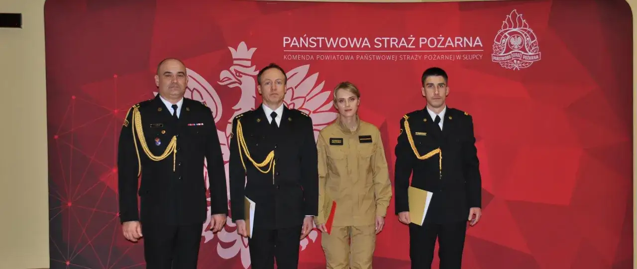 Czterech funkcjonariuszy Państwowej Straży Pożarnej stoi przed czerwonym tłem. Dyskutują o tym, jakie stopnie w PSP można osiągnąć po ilu latach służby.