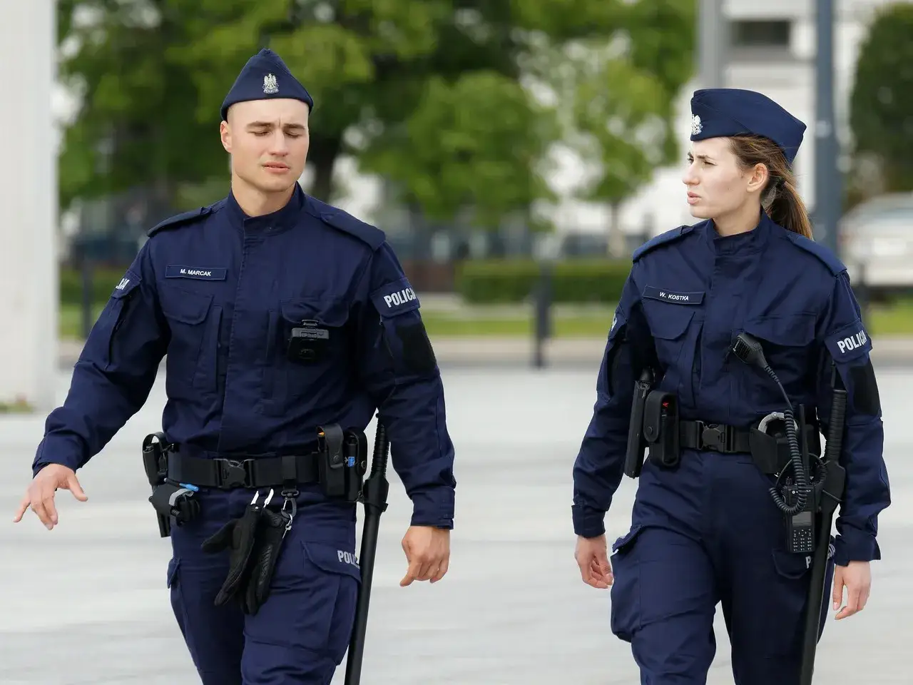 Policjanci w pełnym rynsztunku, z widocznym wyposażeniem: pałka, kajdanki, kabura z pistoletem.