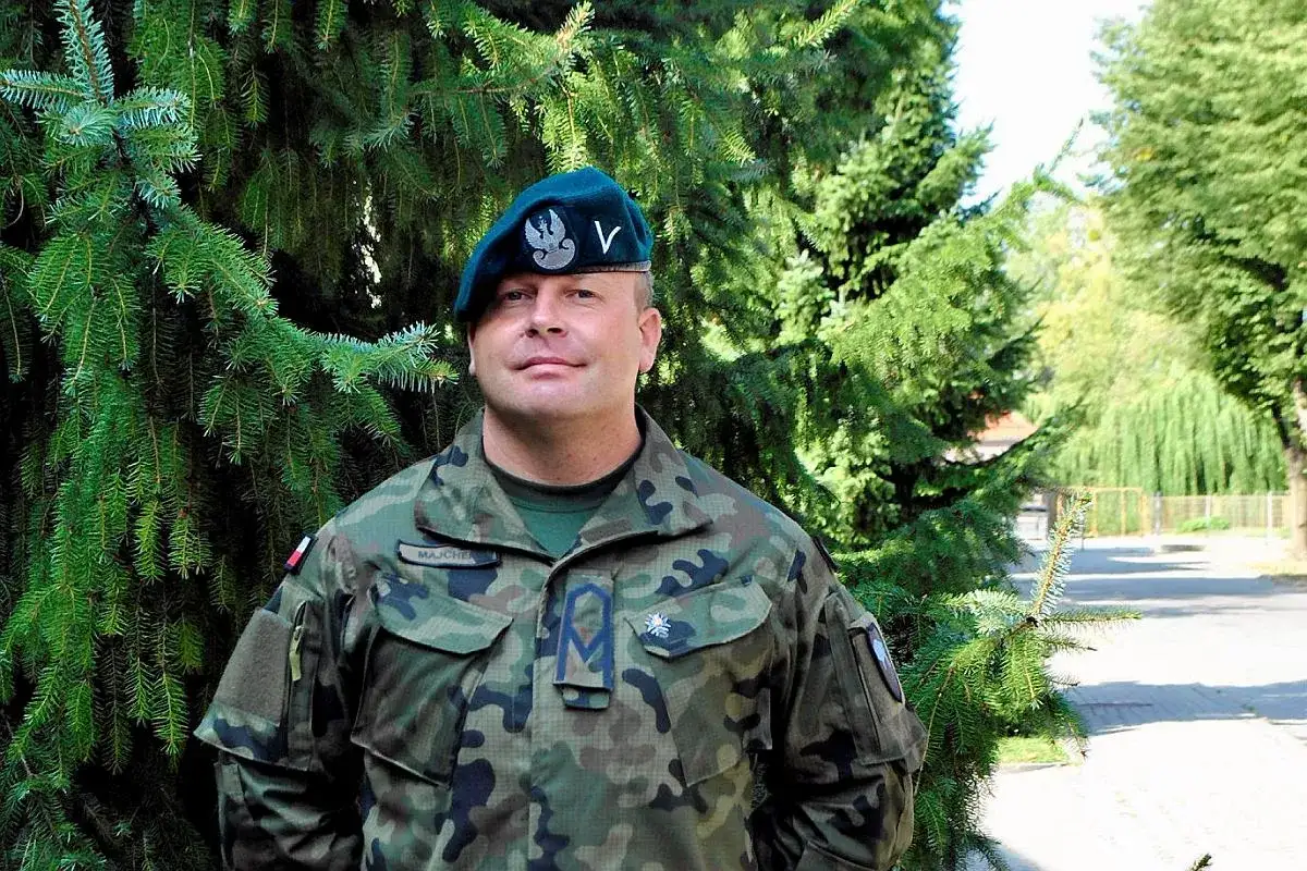 Sierżant w wojsku, w mundurze moro i zielonym beretcie z orłem, stoi na tle drzew.