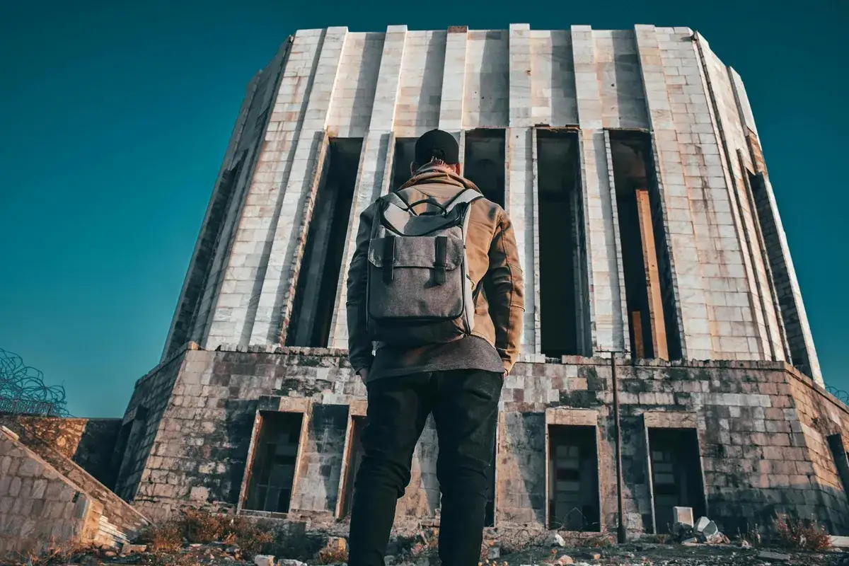 Osoba z plecakiem przed monumentalnym, opuszczonym budynkiem. To przygoda z chodzenia po opuszczonych budynkach, odkrywanie zapomnianych miejsc.
