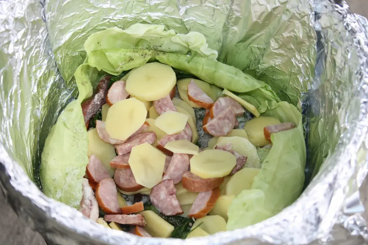 Warzywa na ognisko: pokrojone ziemniaki, kiełbasa i kapusta zawinięte w folię aluminiową, gotowe do pieczenia.