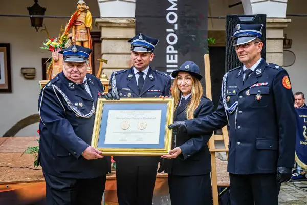 Członkowie OSP otrzymują odznaczenie. W tle drewniana figura i baner.